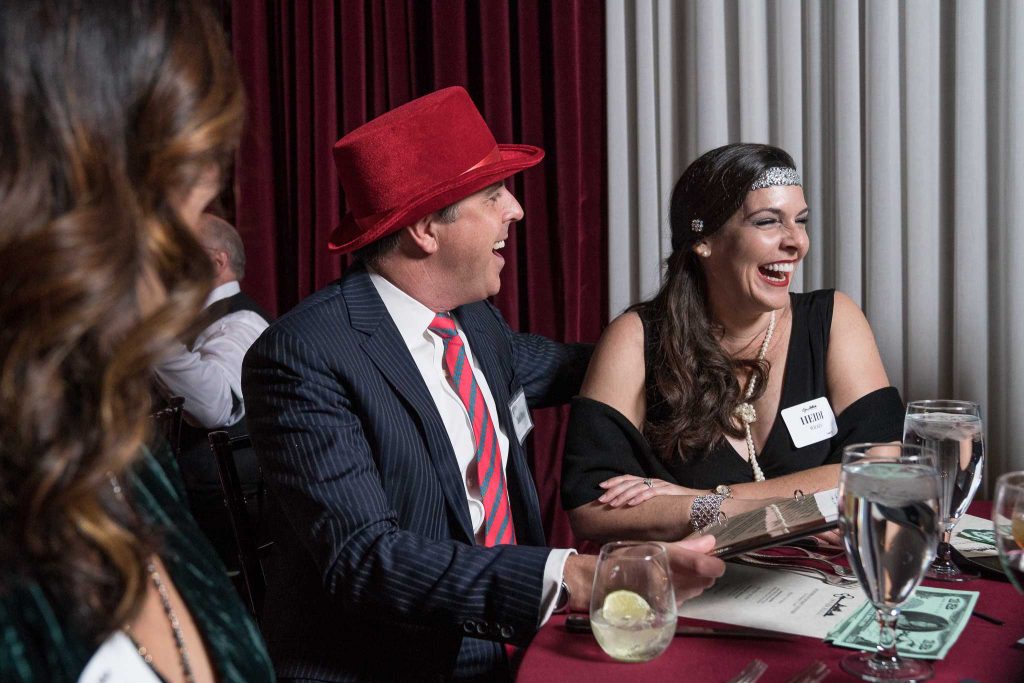 A sharply-dressed man in a red hat and a woman in formal attire laugh together during an immersive theater event